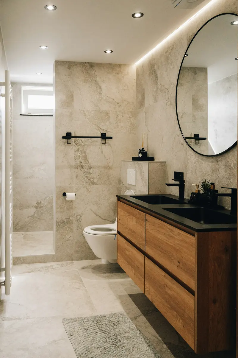 Baño en tono piedra con mueble de madera y espejo redondo